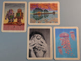 Four illustrated cards arranged on a gray surface. The cards feature surreal artwork: two cards show colorful landscapes and figures, one card is a black-and-white portrait of hands covering a face, and one card shows pink jellyfish on a blue background.